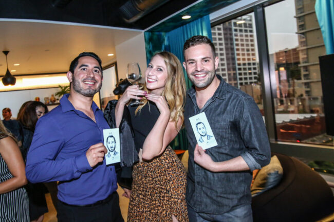 Three guests at an event smiling and holding up their hand-drawn portrait sketches, with a city view in the background.